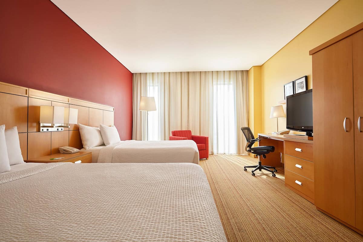 Habitación de hotel moderna con dos camas y área de trabajo, con iluminación natural y decoración cálida.