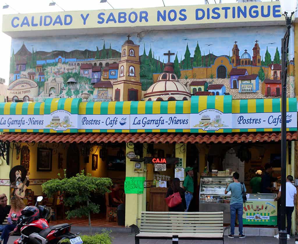 Fachada colorida de La Garrafa-Nieves en León, con cartel de Postres y Café, y ambiente animado en la calle.