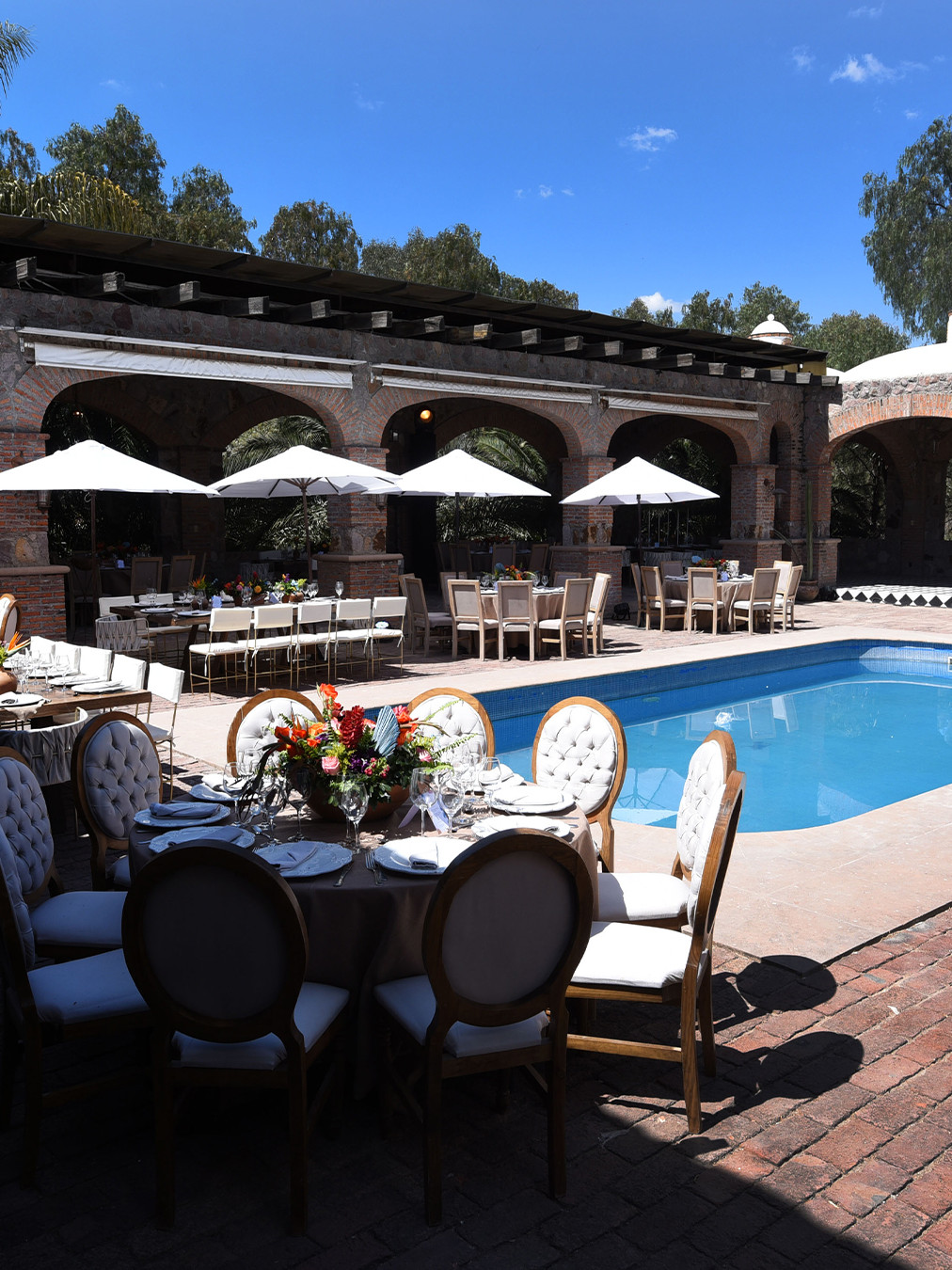 Mesa decorada con flores y mantelería en un espacio con piscina y sombra, ideal para disfrutar en León, México.
