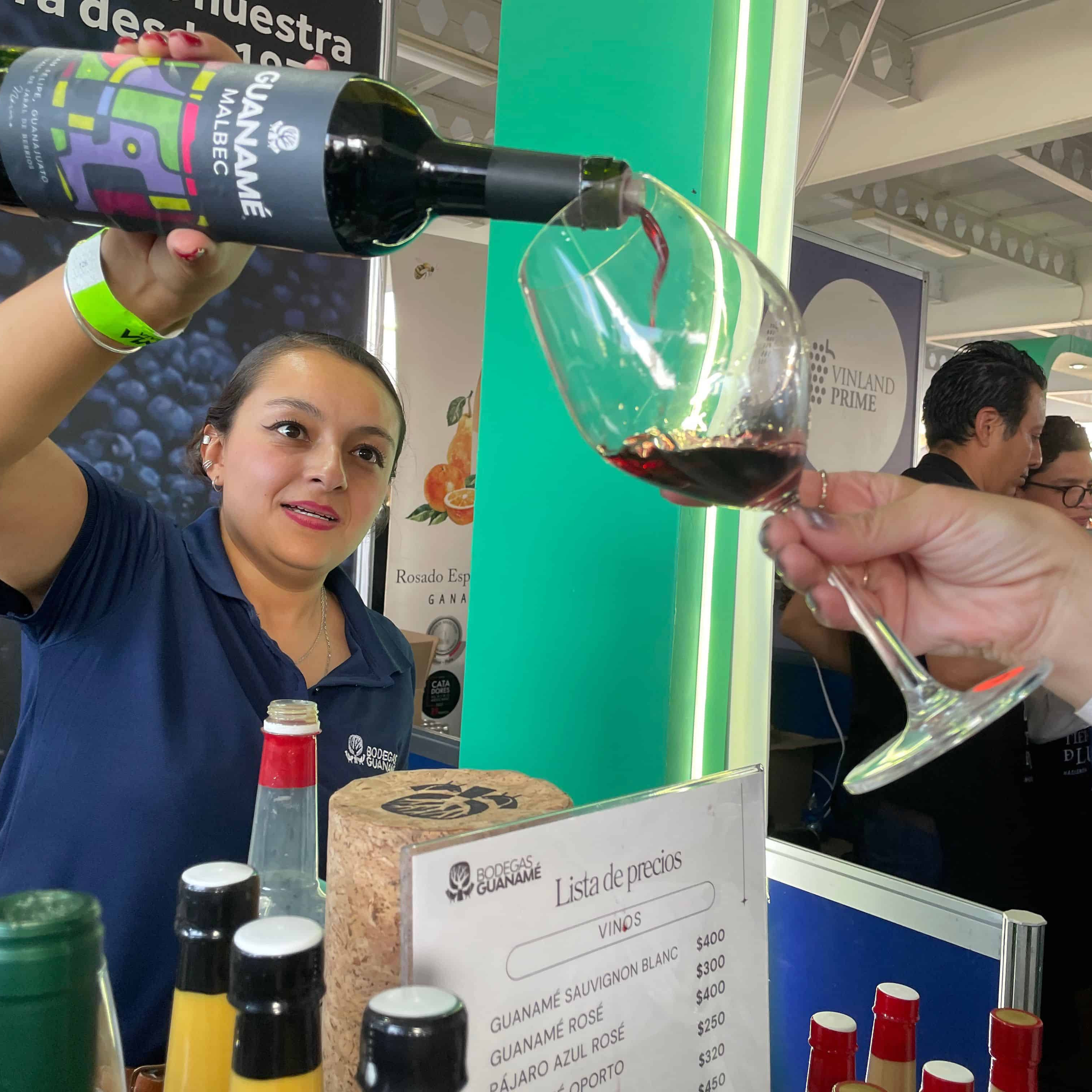 Vendedora sirviendo vino tinto en feria gastronómica en León, México, con ambiente animado y degustación de vinos locales.