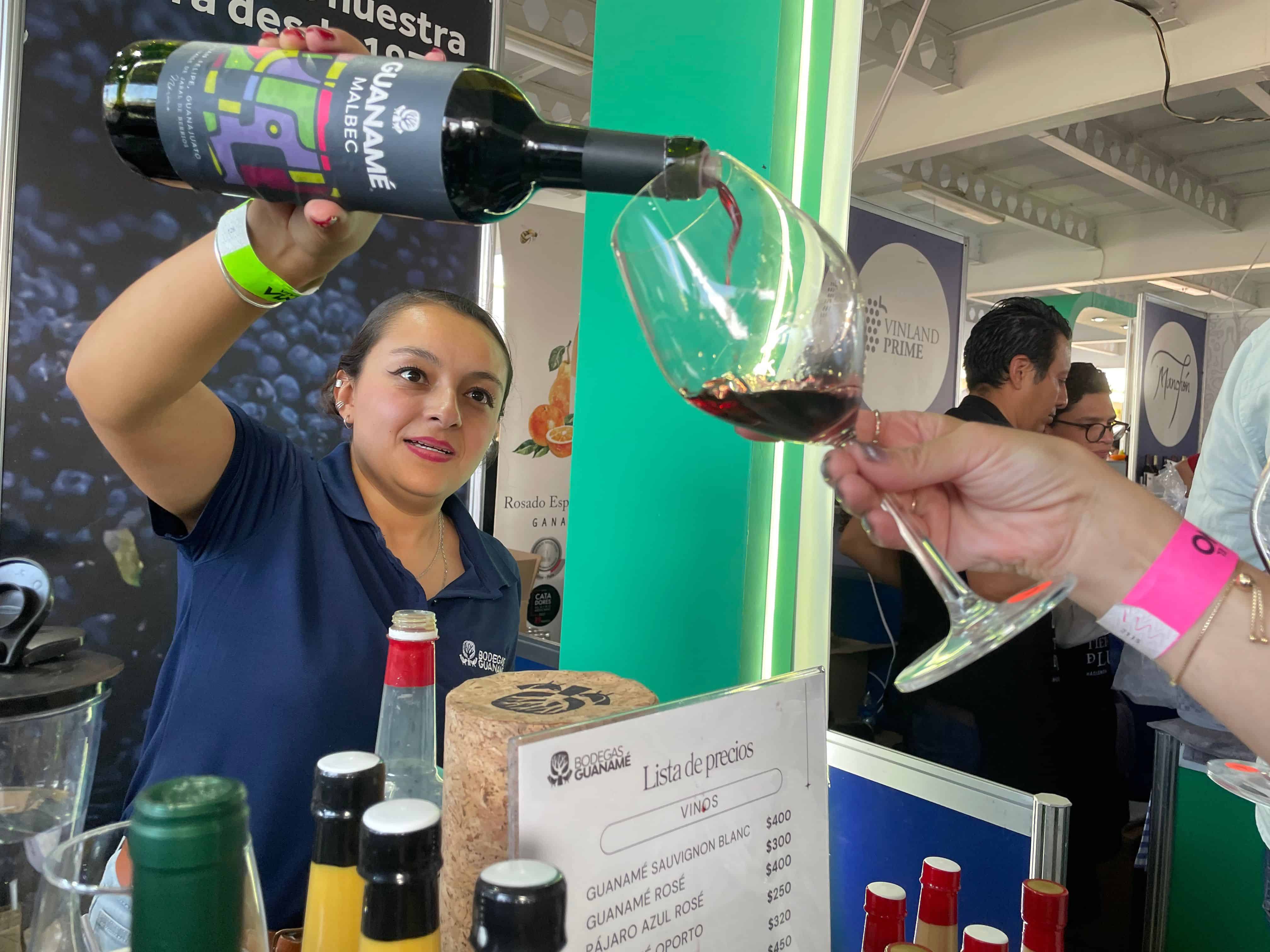 Vendedora sirviendo vino tinto en feria gastronómica en León, México, con ambiente animado y degustación de vinos locales.