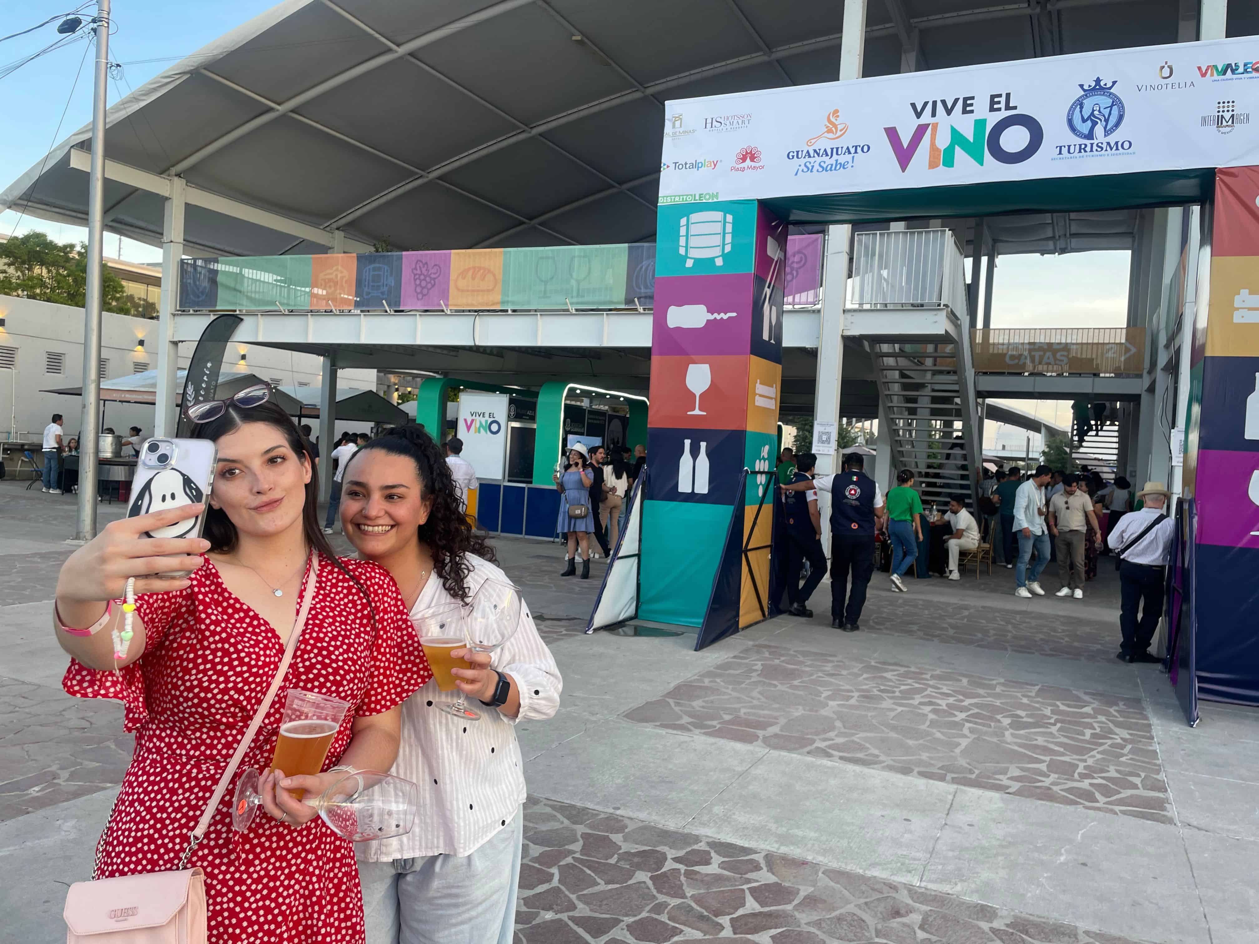 Mujeres en evento de vino en León, México, tomando selfies y disfrutando de la feria de vino y gastronomía.