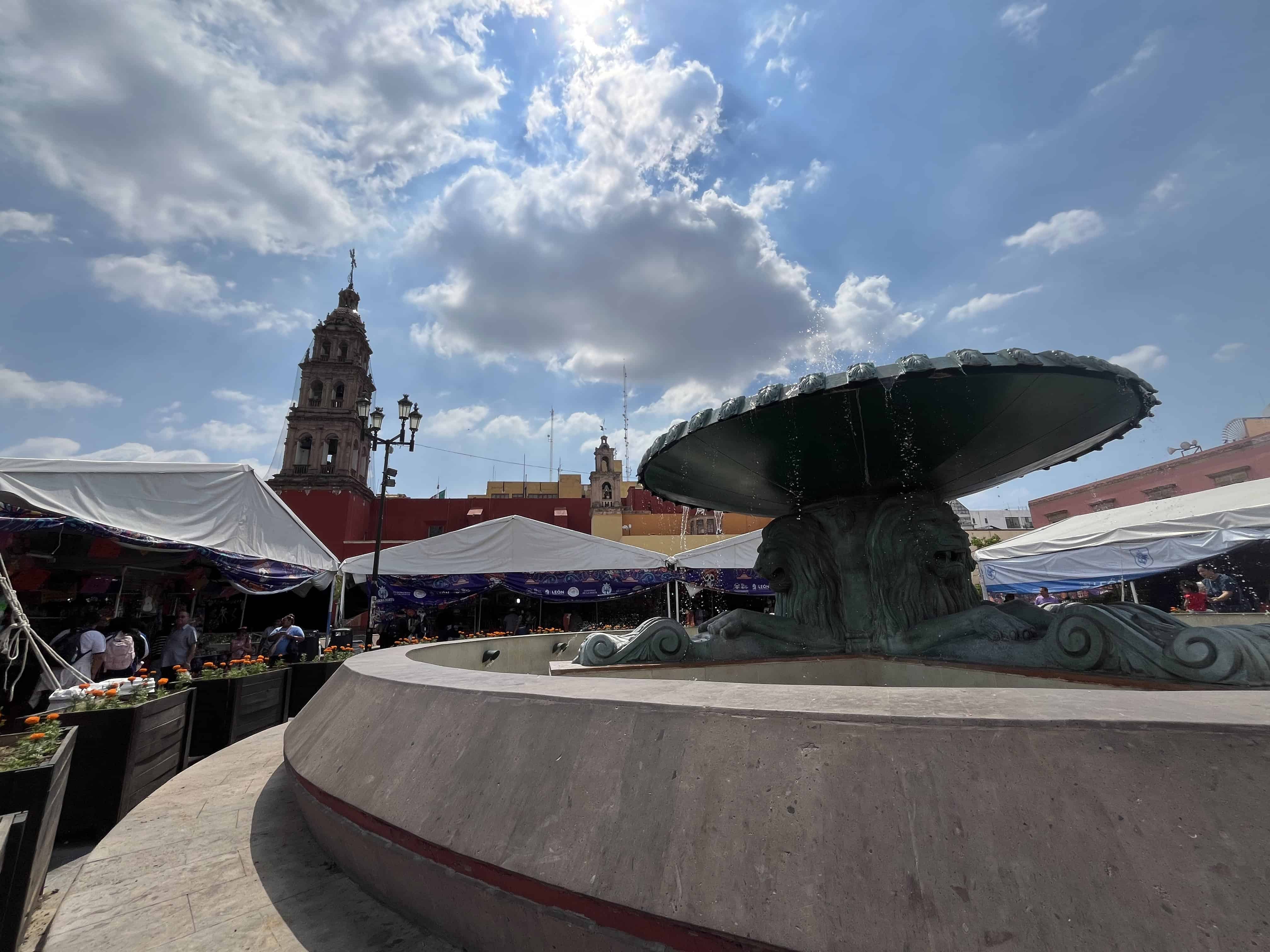 Fuente de la Plaza Principal en Le&oacute;n, Guanajuato, rodeada de puestos de artesan&iacute;as y edificios hist&oacute;ricos en un d&iacute;a soleado.