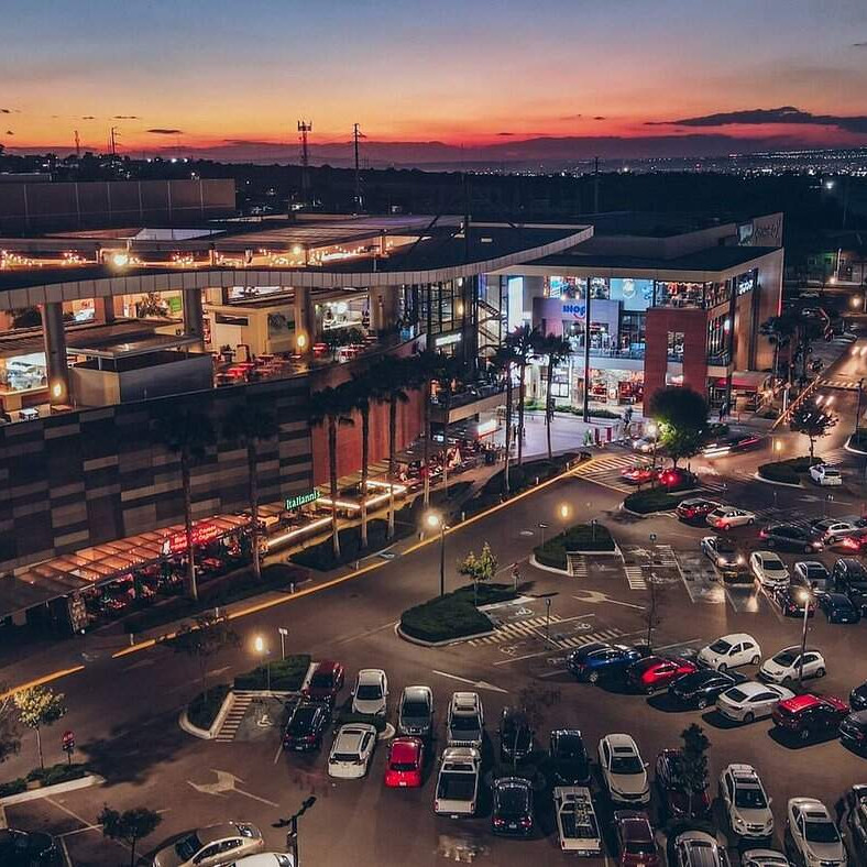 Centro comercial en León, México, iluminado al atardecer con estacionamiento lleno de autos.