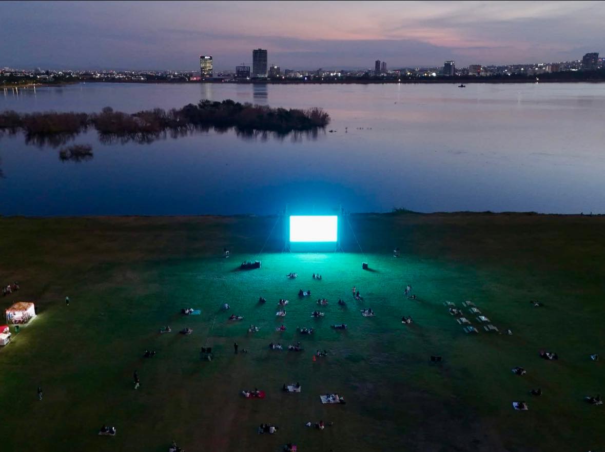 Proyecci&oacute;n de pel&iacute;cula en pantalla gigante junto al r&iacute;o, con espectadores disfrutando en un entorno natural y urbano.