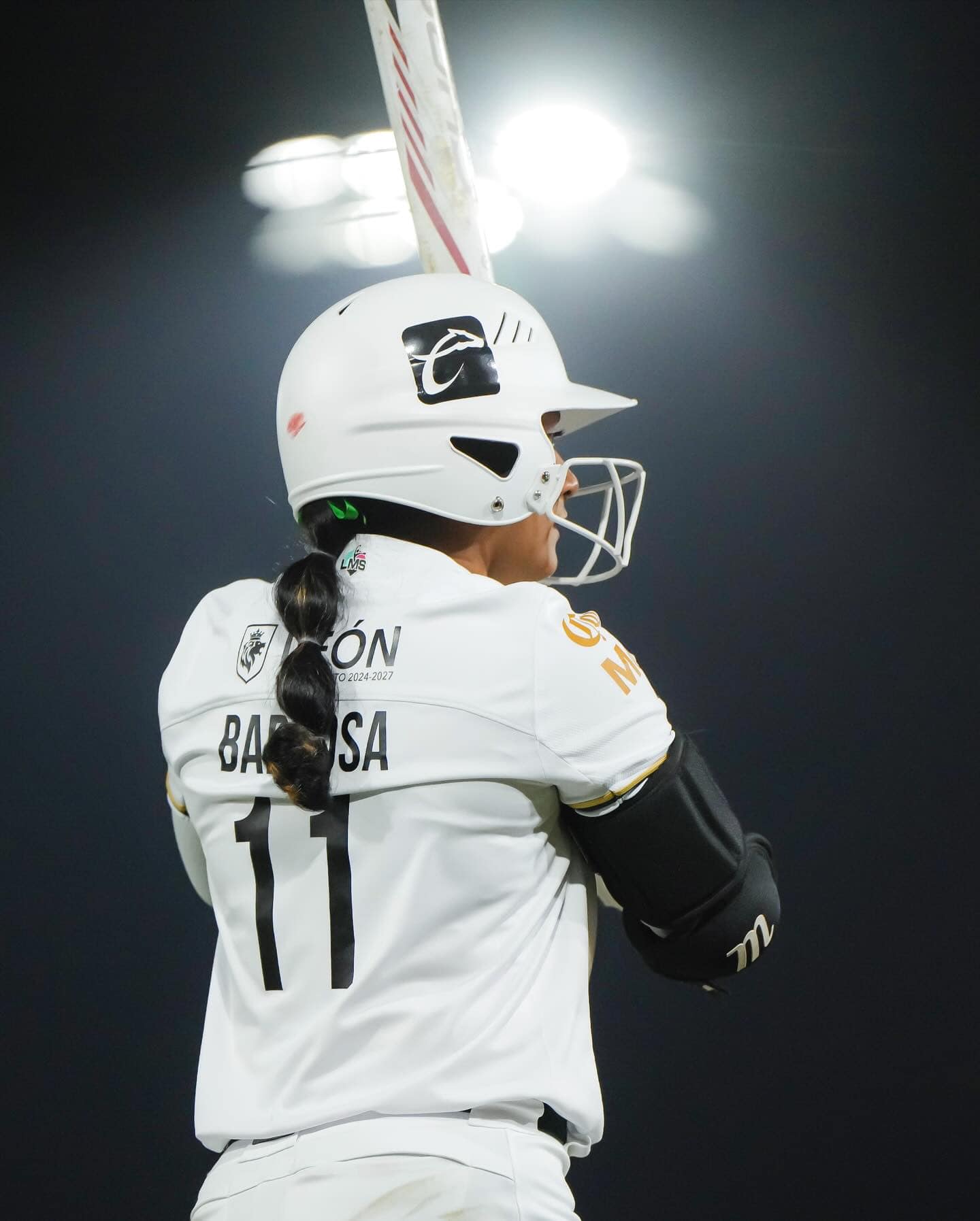 Jugadora de b&eacute;isbol femenina en uniforme blanco, con casco y bate, lista para batear en un partido emocionante.