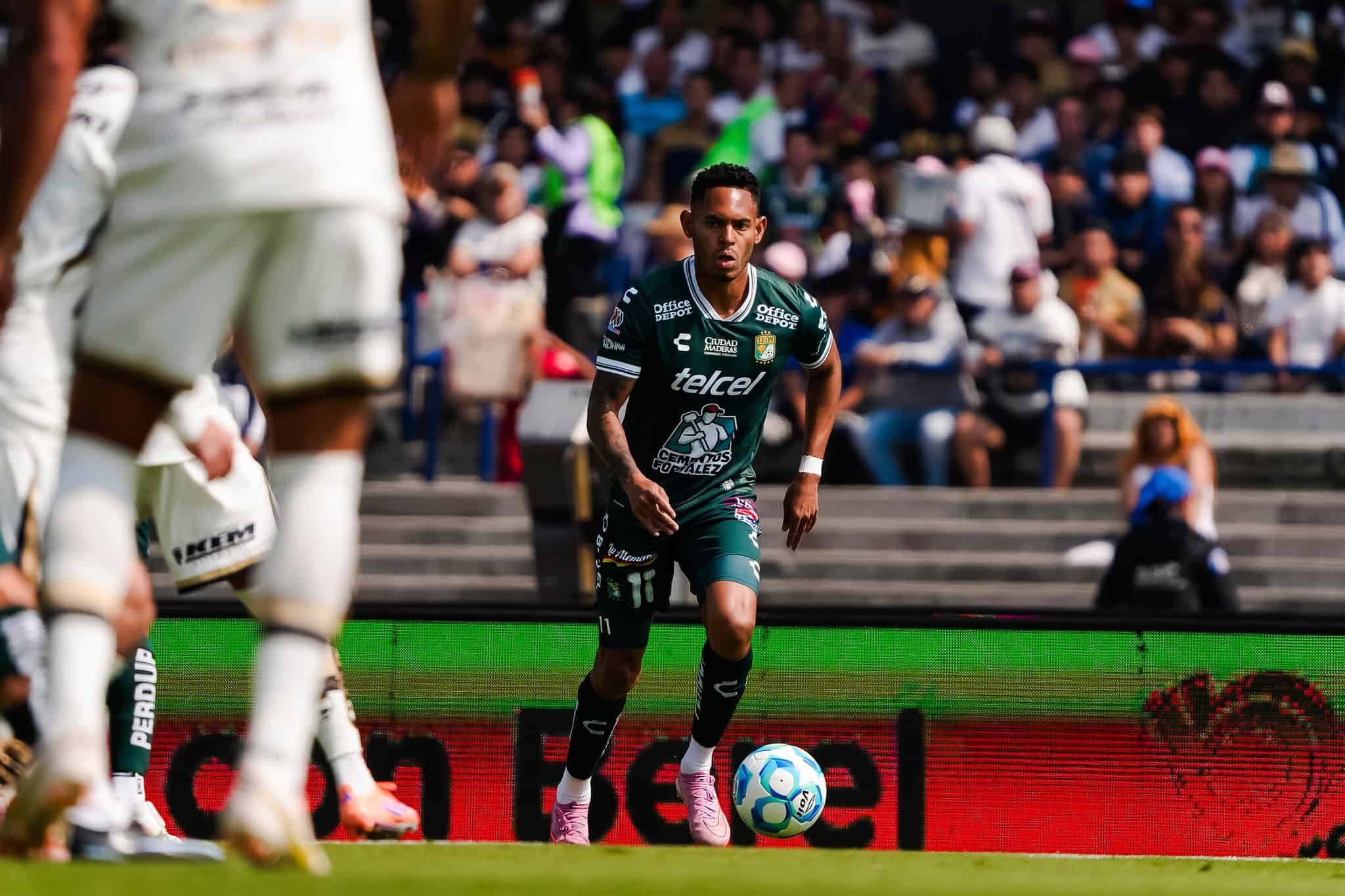 Jugador de f&uacute;tbol en acci&oacute;n durante un partido en Le&oacute;n, con p&uacute;blico en las gradas.