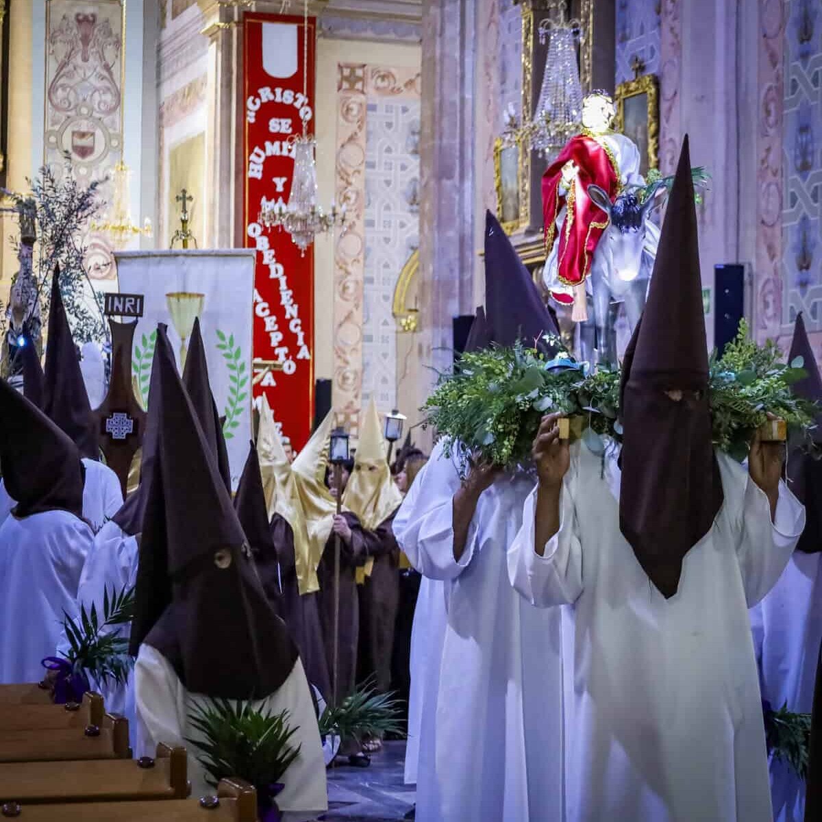 Personas vestidas con trajes típicos y capuchas participan en procesión religiosa en una iglesia histórica.