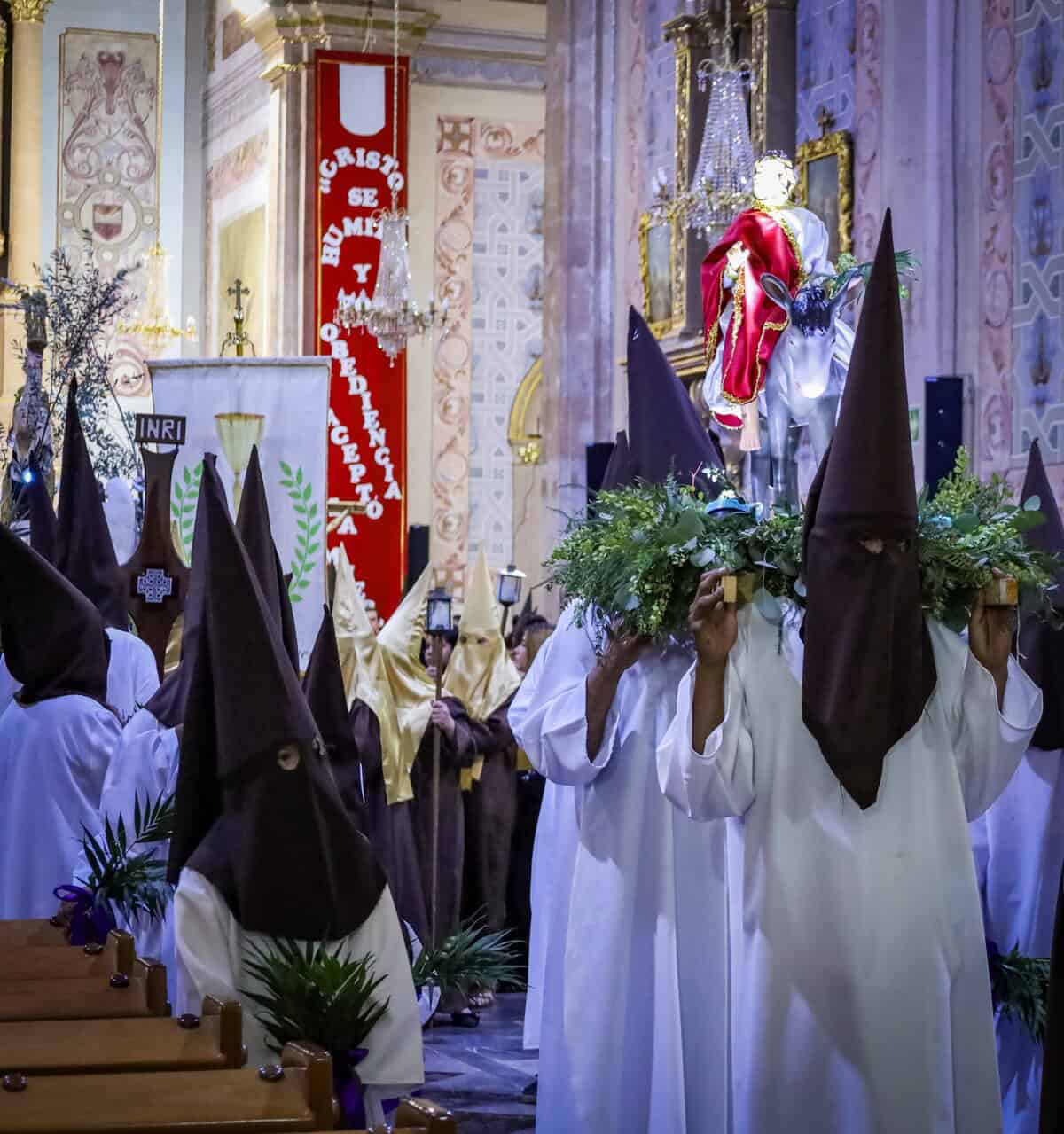 Personas vestidas con trajes típicos y capuchas participan en procesión religiosa en una iglesia histórica.