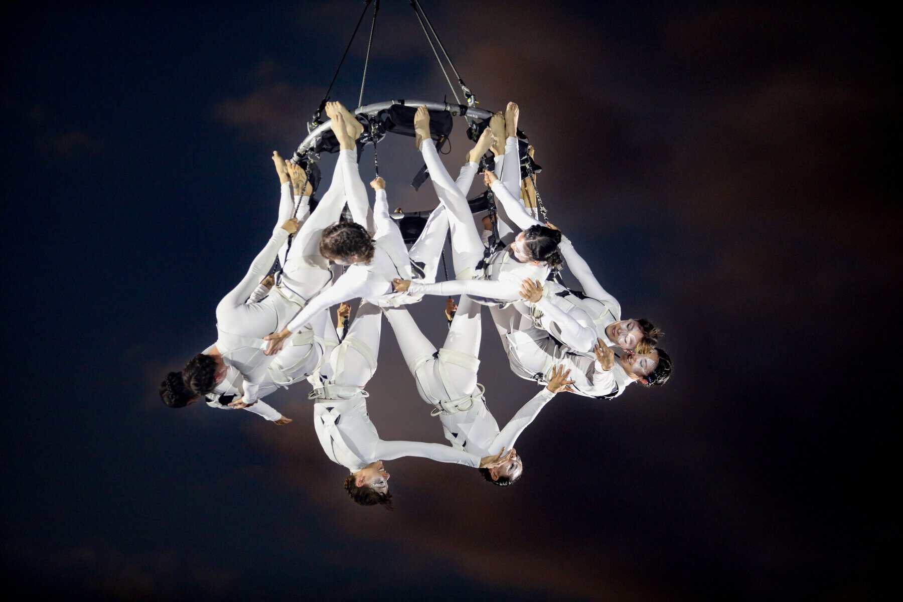 Espect&aacute;culo de circo a&eacute;reo con artistas en trajes blancos realizando acrobacias en el aire.