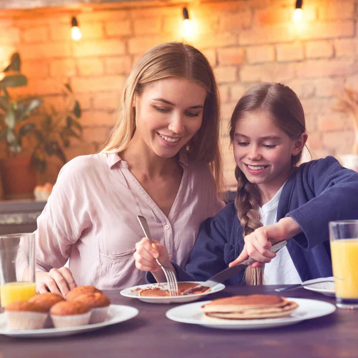 Una madre e hija disfrutan juntas un desayuno con jugo, pan y hotcakes en un ambiente cálido y moderno en León, México.