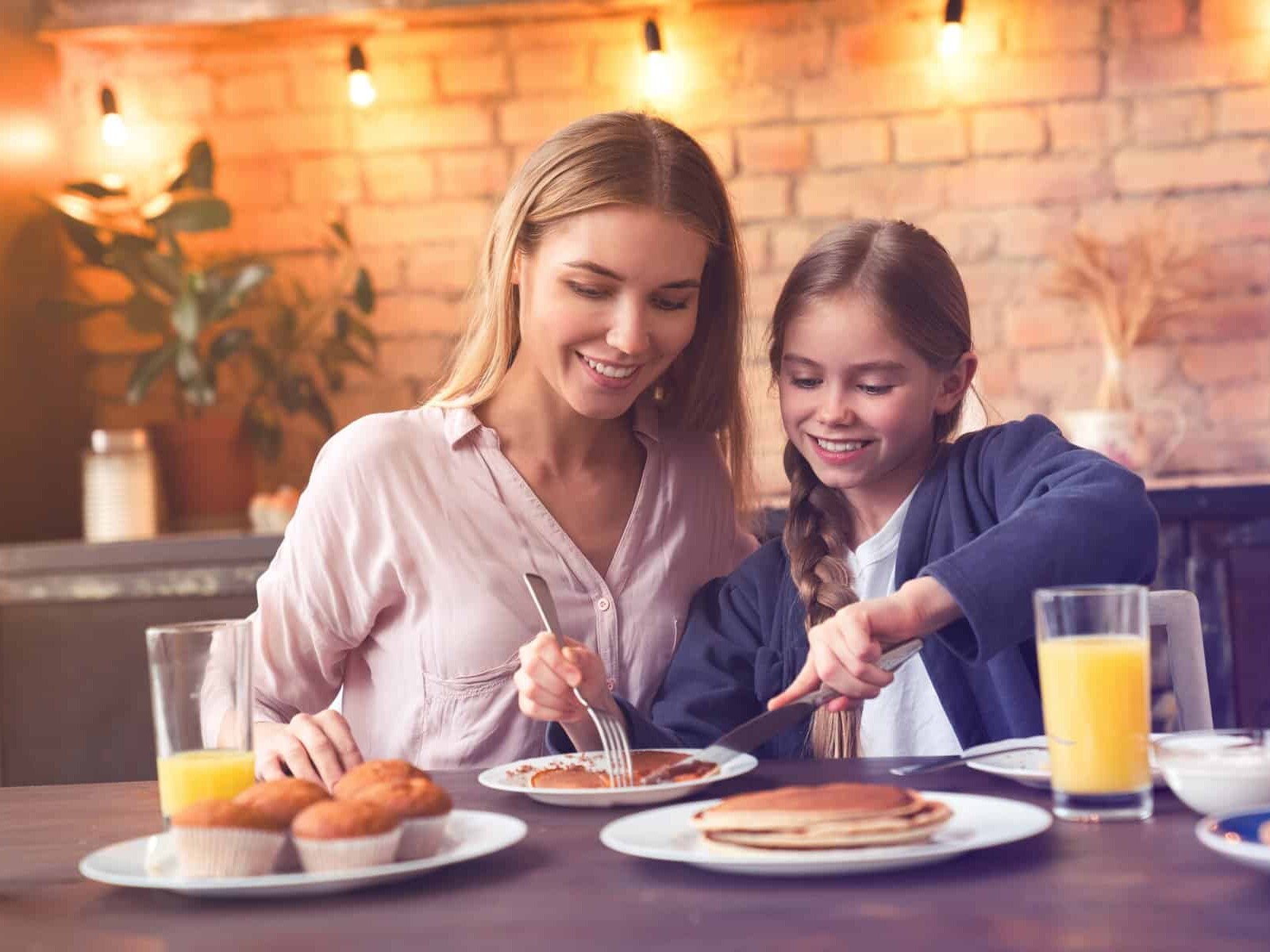 Una madre e hija disfrutan juntas un desayuno con jugo, pan y hotcakes en un ambiente cálido y moderno en León, México.