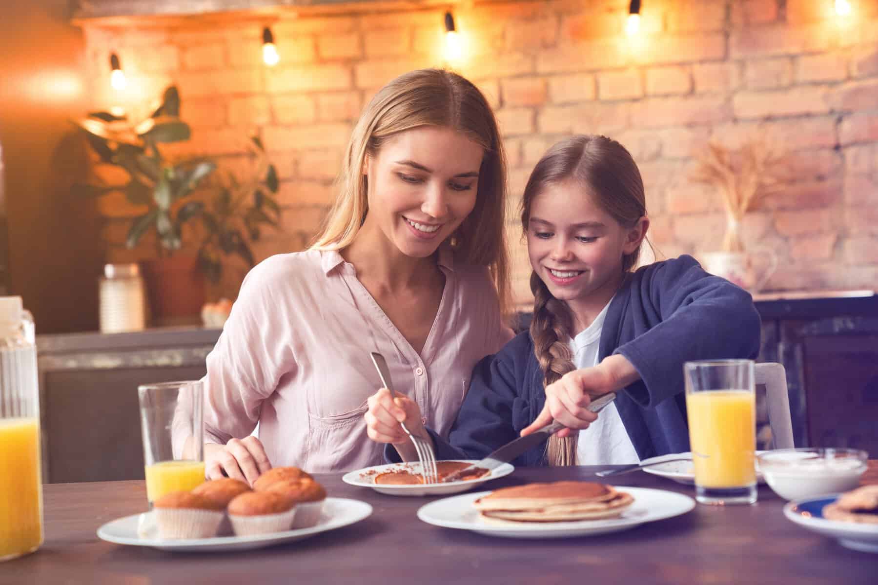 Una madre e hija disfrutan juntas un desayuno con jugo, pan y hotcakes en un ambiente cálido y moderno en León, México.