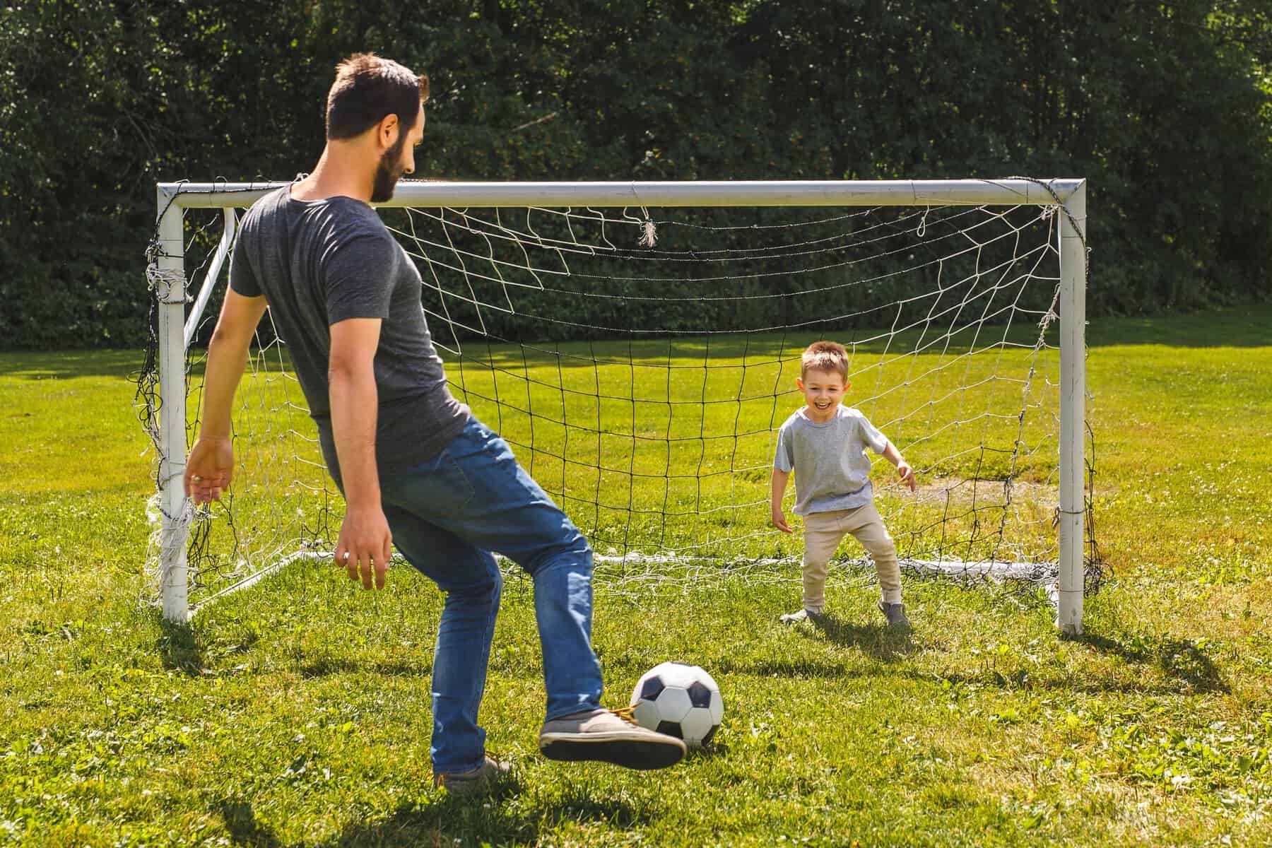 Un adulto y un ni&ntilde;o jugando f&uacute;tbol en un campo al aire libre, con un arco y bal&oacute;n en un d&iacute;a soleado.