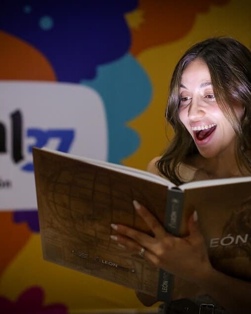 Mujer emocionada leyendo en la feria del libro en León, con fondo del evento “León 450: Dejando Huella en la Historia”.