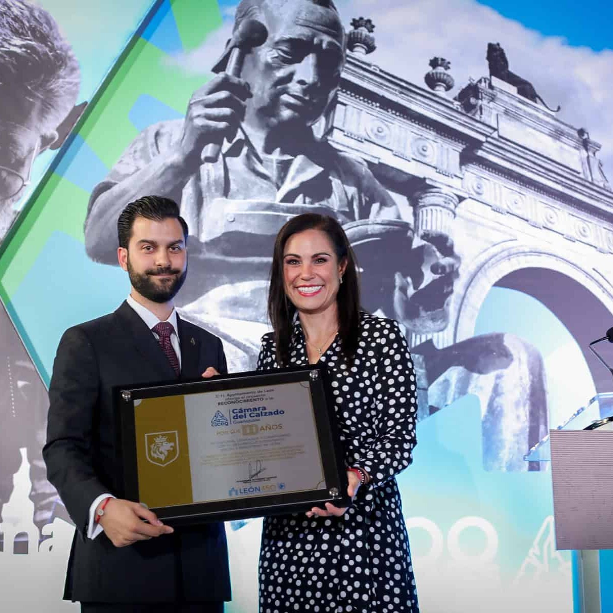 Dos personas en ceremonia de premiación en León, con monumentos históricos y arte en el fondo, celebrando logros culturales y.