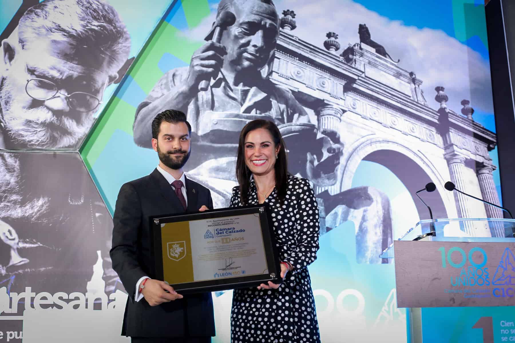 Dos personas en ceremonia de premiación en León, con monumentos históricos y arte en el fondo, celebrando logros culturales y.