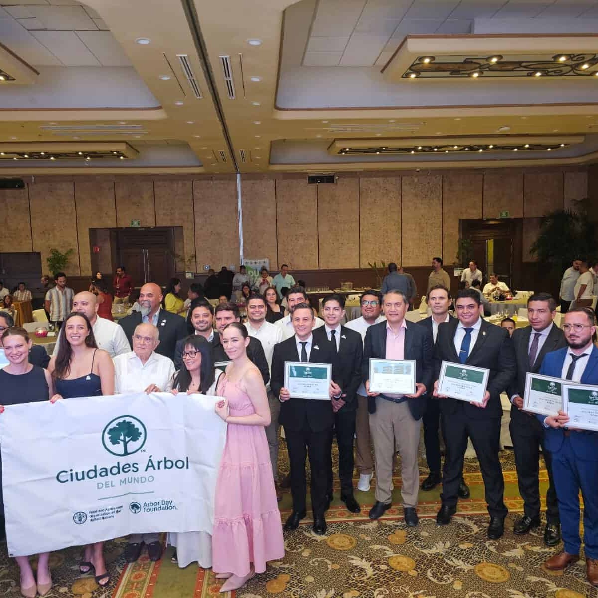 Grupo de personas en evento de Ciudades Árbol del Mundo, sosteniendo un banner y certificados en un salón de conferencias.