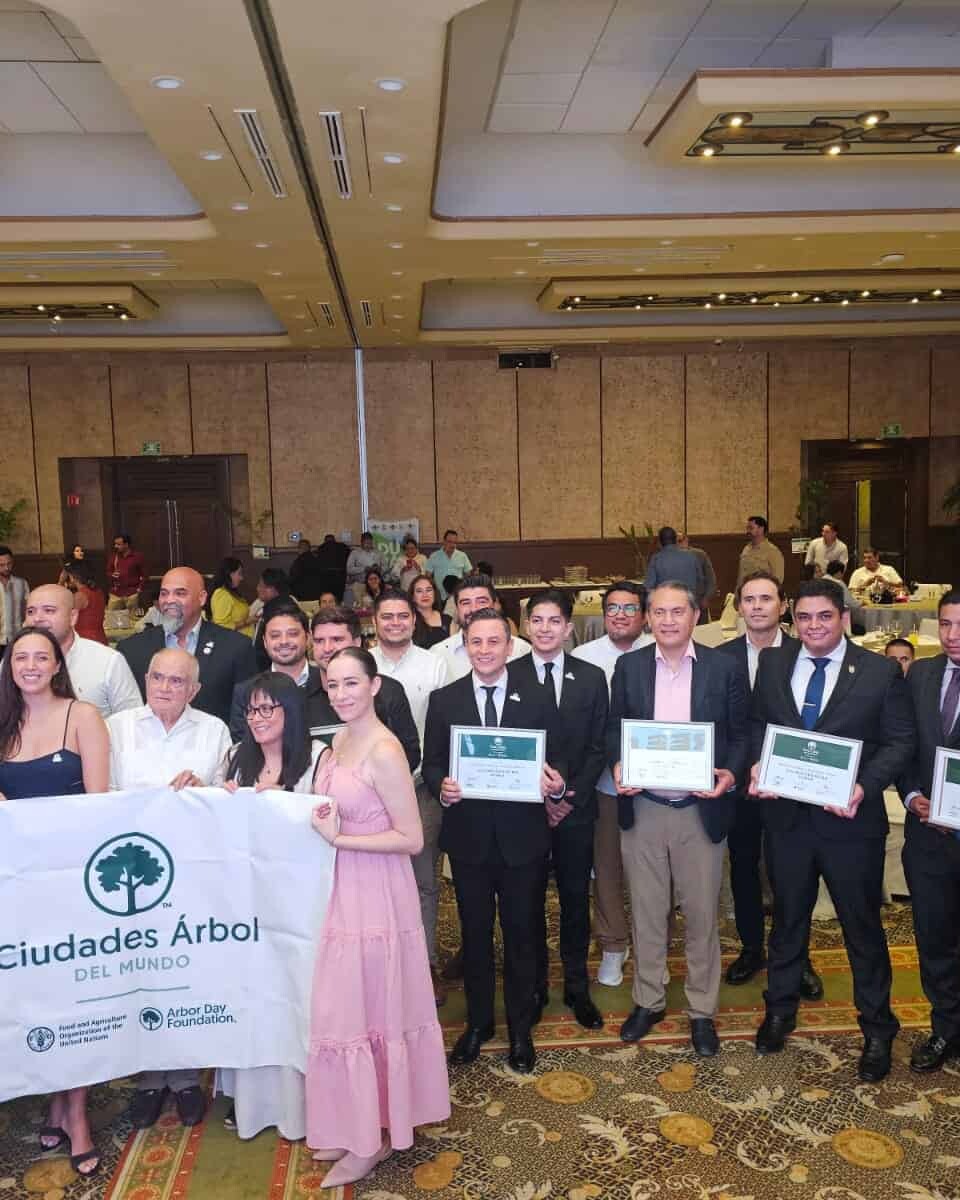 Grupo de personas en evento de Ciudades Árbol del Mundo, sosteniendo un banner y certificados en un salón de conferencias.