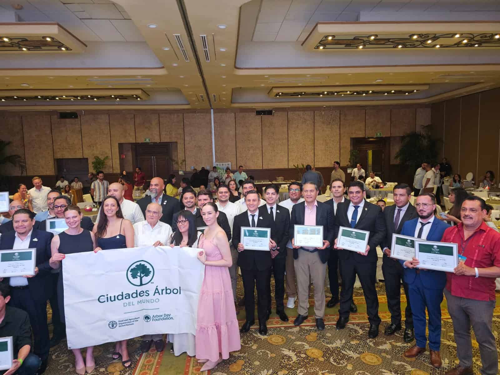Grupo de personas en evento de Ciudades &Aacute;rbol del Mundo, sosteniendo un banner y certificados en un sal&oacute;n de conferencias.