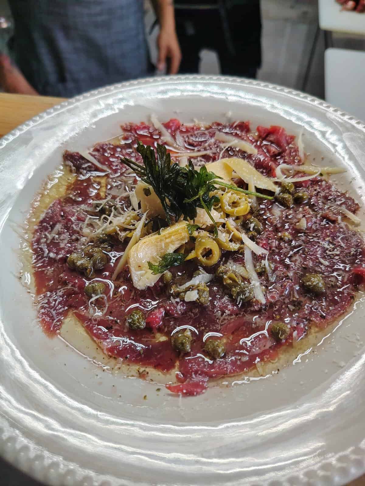 Carpaccio de res con aderezos frescos, aceitunas, alcaparras y hierbas aromáticas en un plato elegante.