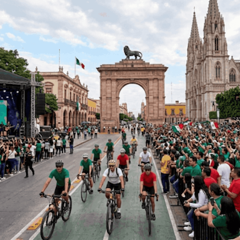 Celebración en León, México, con público y ciclistas en la calle frente a la catedral y el Arco de la Independencia.