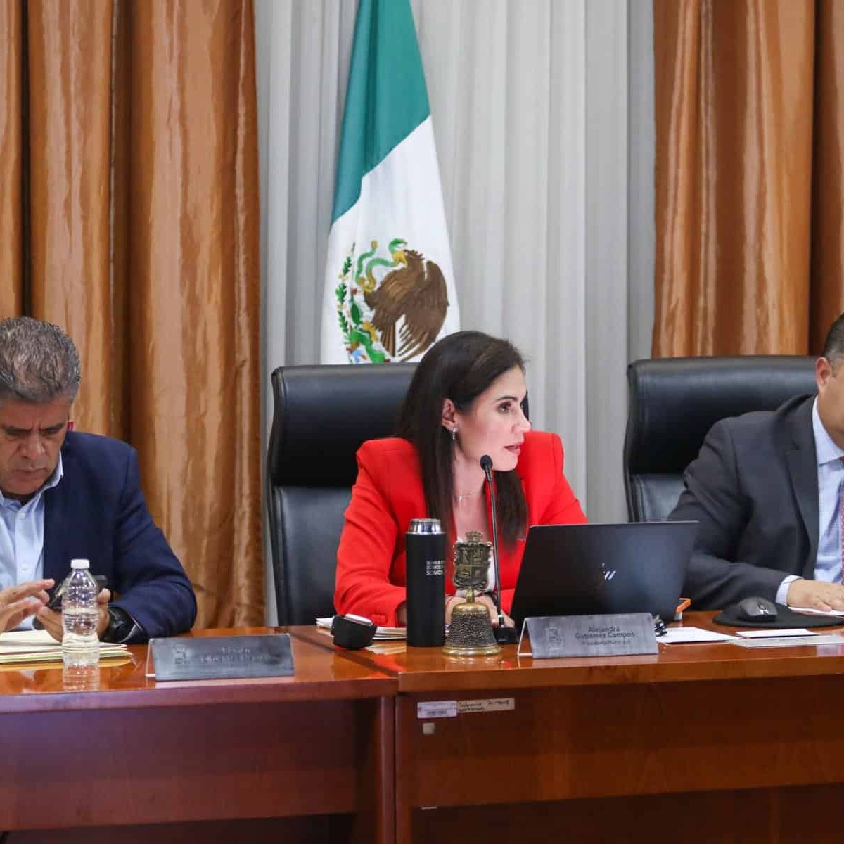Tres funcionarios en una reunión oficial en el Palacio de Gobierno de León, con la bandera de México al fondo.