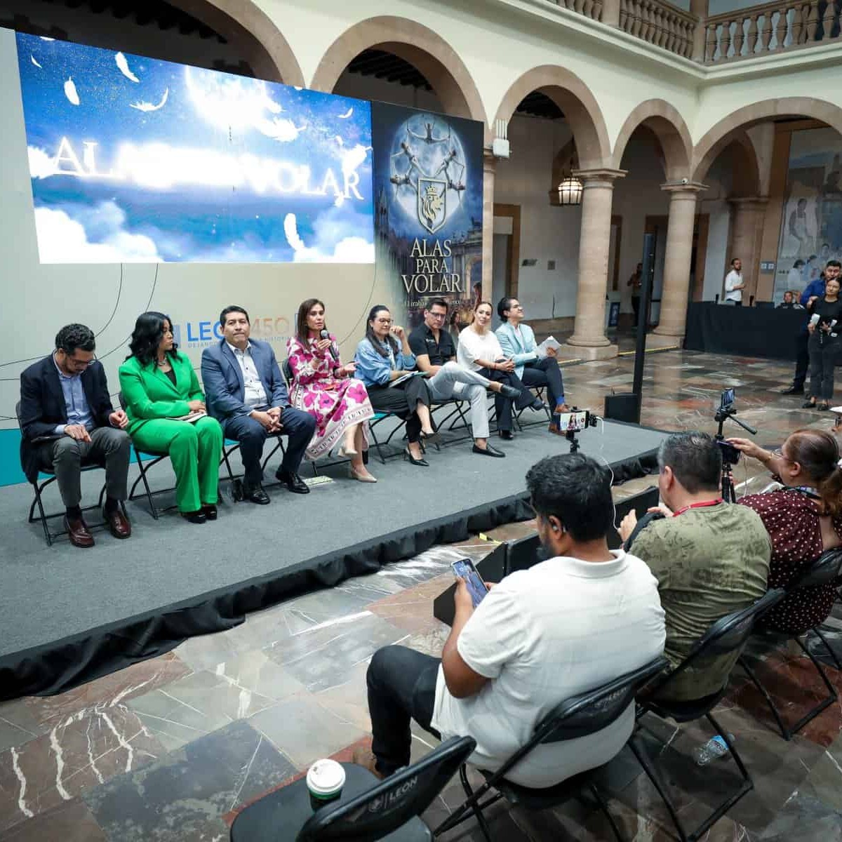 Panel de expertos en un evento en León, México, con audiencia y pantalla grande en un espacio histórico y elegante.
