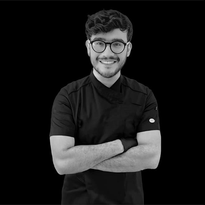Joven chef sonriendo con gafas y uniforme negro, de pie con brazos cruzados, fondo negro.