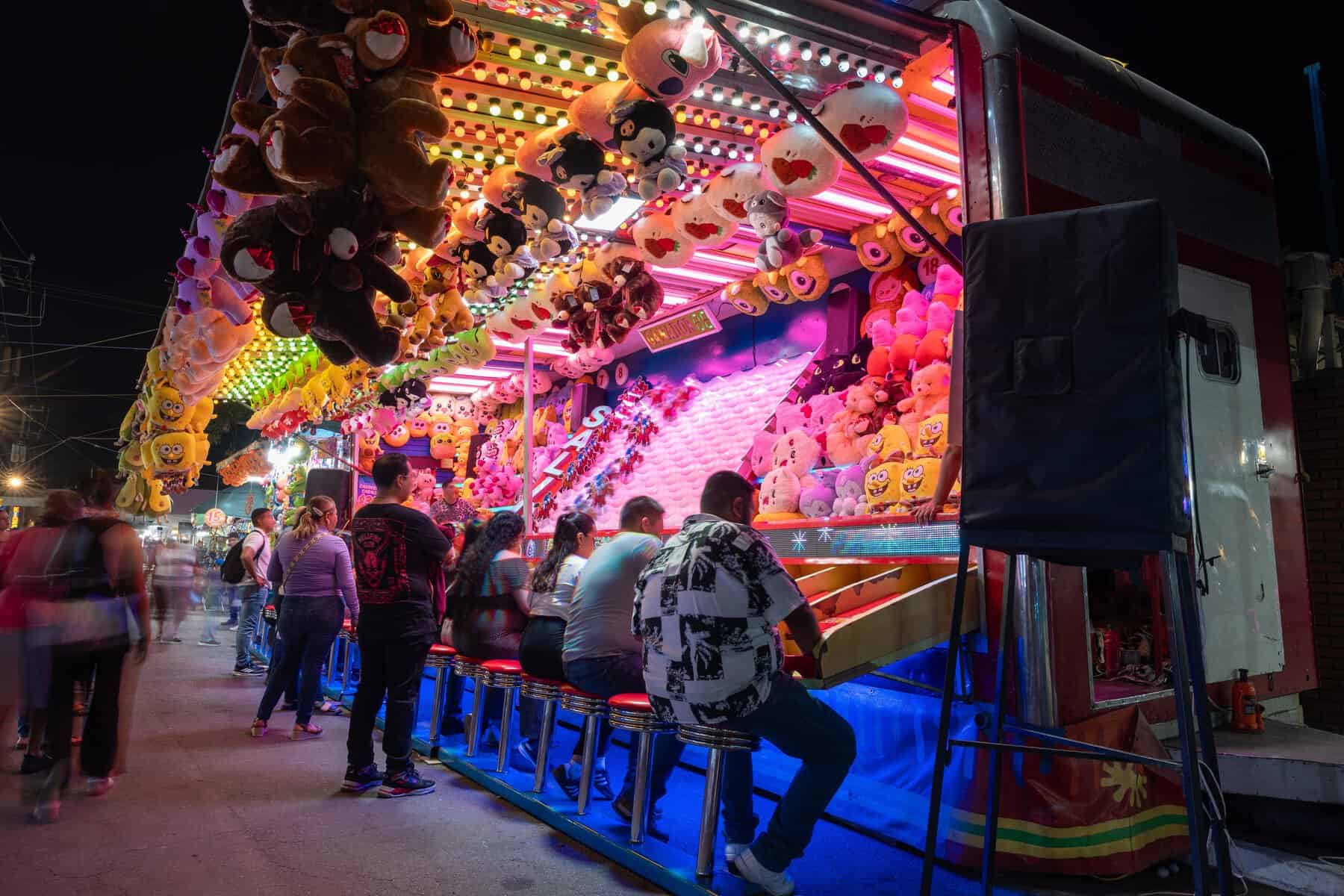 Juegos de feria en León Guanajuato con luces y peluches colgando, personas disfrutando en la noche.