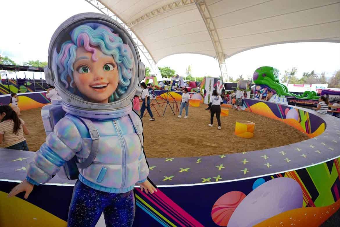 Niña con disfraz de astronauta en parque de diversiones en León, Guanajuato, México, con atracciones y público disfrutando.