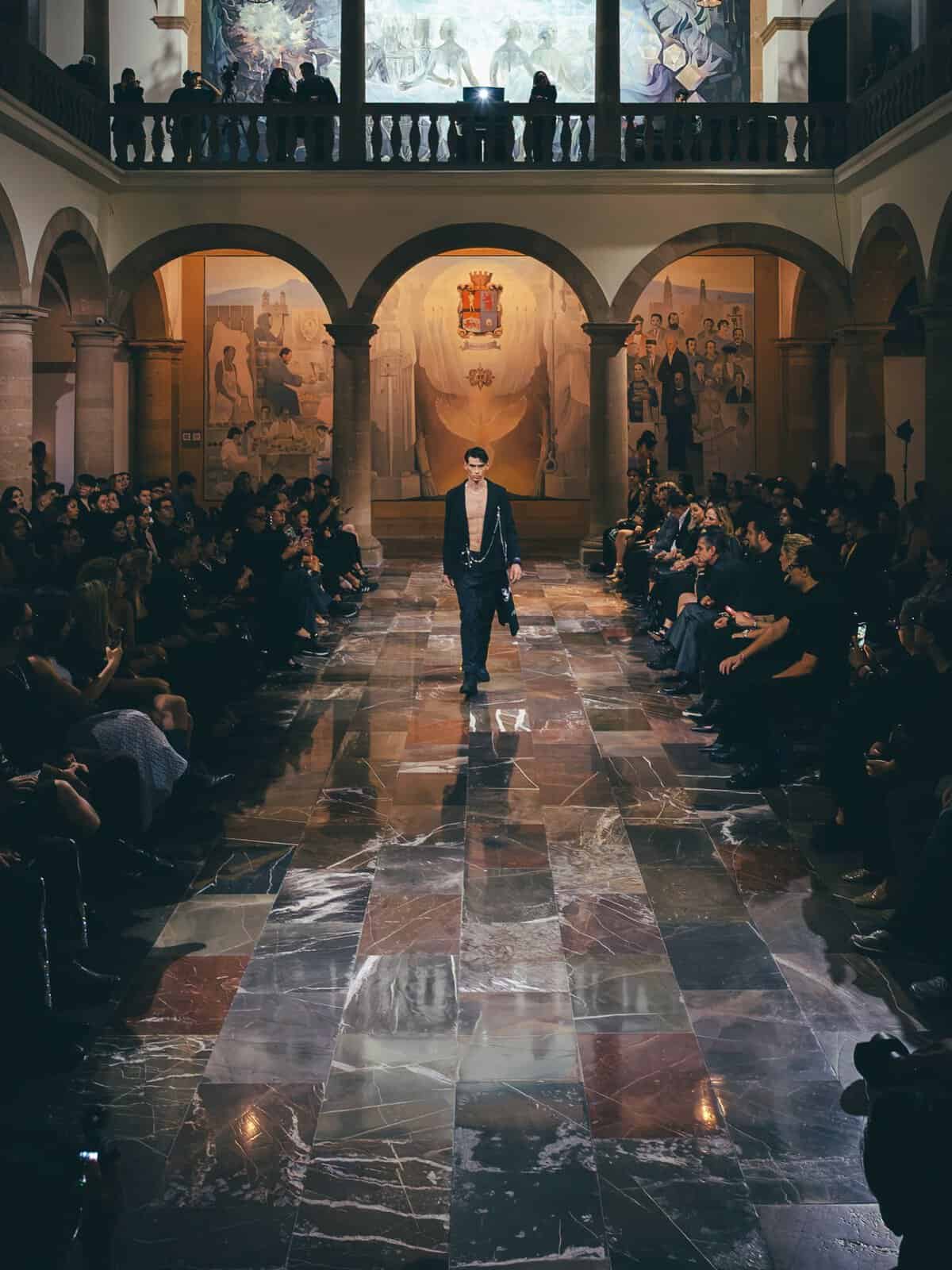 Modelo caminando en pasarela durante evento de moda en León, Guanajuato, México, con público y arte en el fondo.