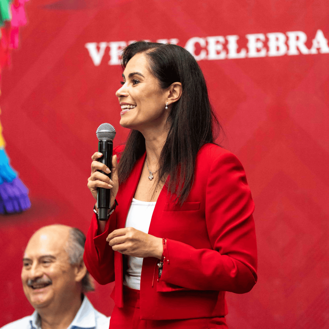 Mujer sonriente en evento cultural en León, Guanajuato, con micrófono y vestimenta roja, promoviendo tradiciones mexicanas.