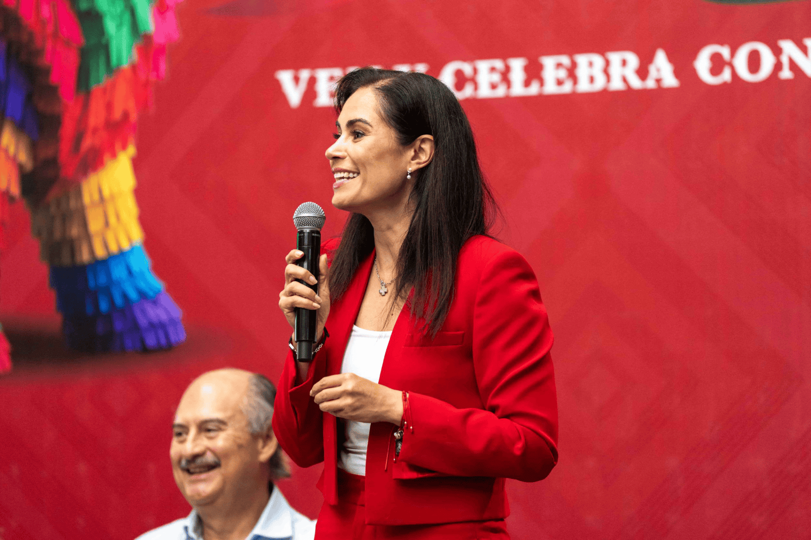 Mujer sonriente en evento cultural en León, Guanajuato, con micrófono y vestimenta roja, promoviendo tradiciones mexicanas.