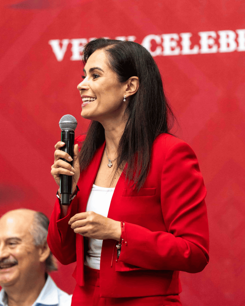 Mujer sonriente en evento cultural en León, Guanajuato, con micrófono y vestimenta roja, promoviendo tradiciones mexicanas.