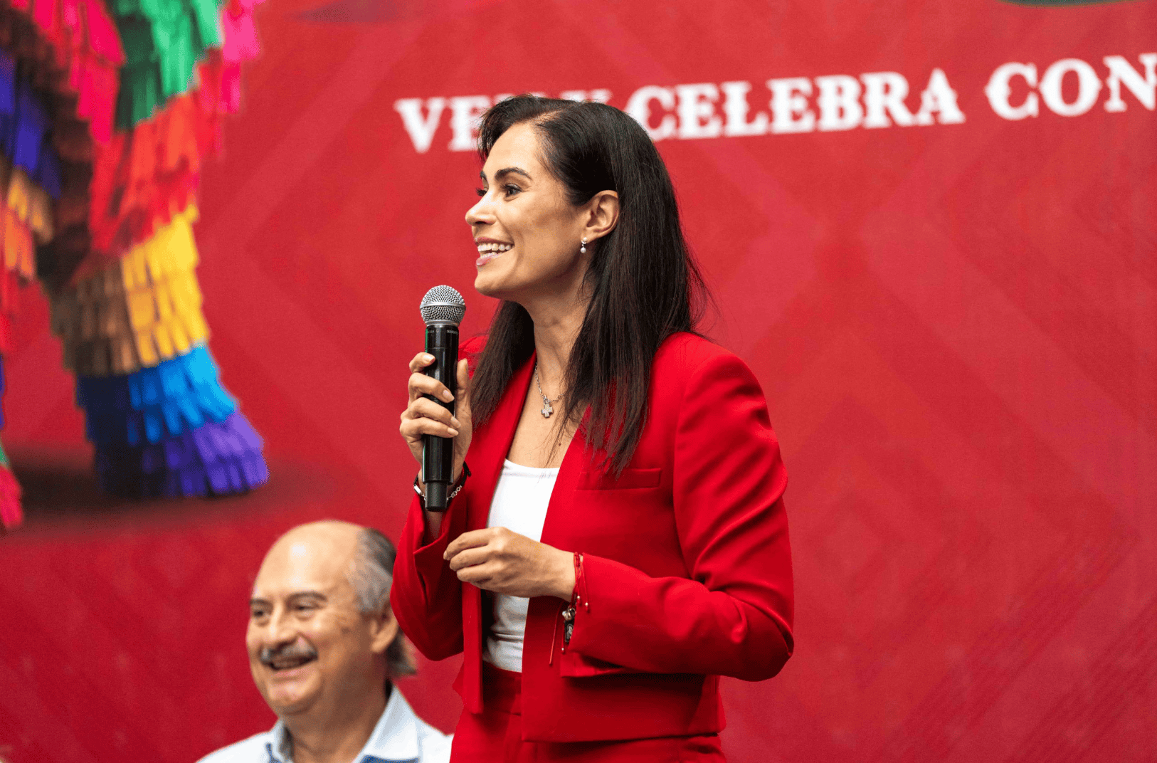 Mujer sonriente en evento cultural en León, Guanajuato, con micrófono y vestimenta roja, promoviendo tradiciones mexicanas.