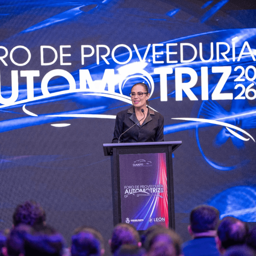 Mujer hablando en foro automotriz en León, Guanajuato, México, con público presente y pantalla digital de fondo.