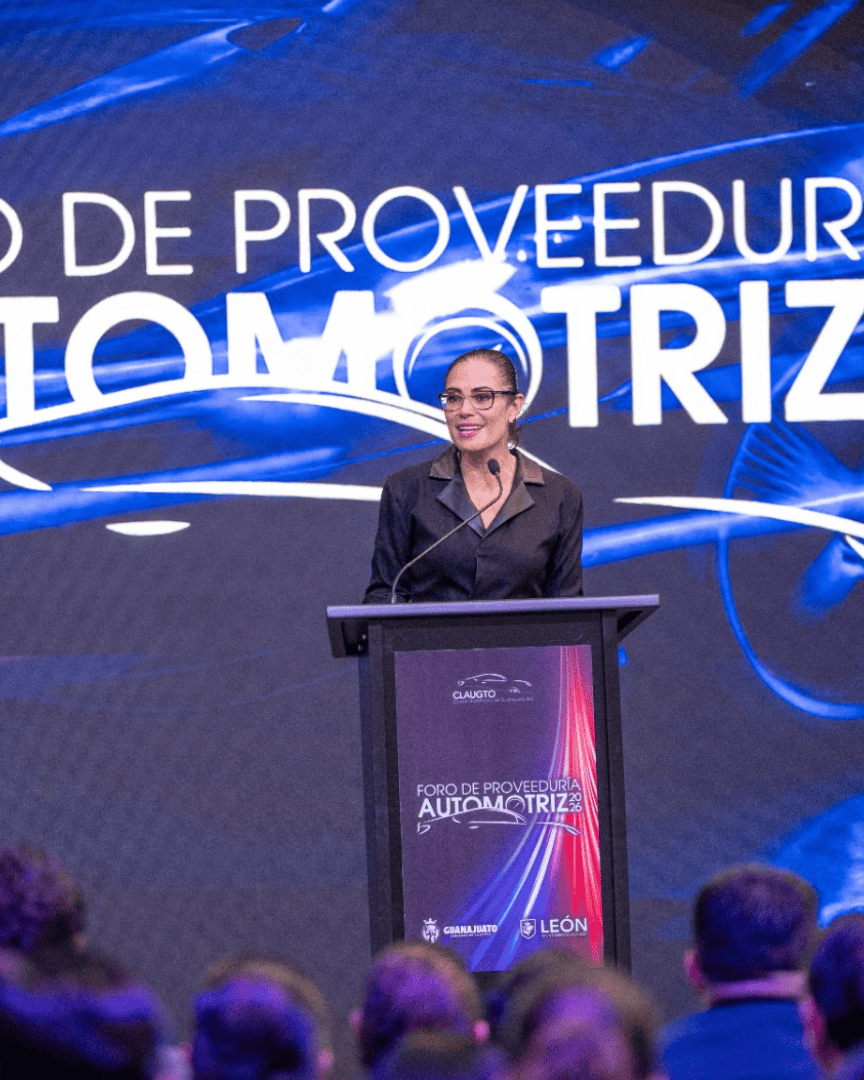 Mujer hablando en foro automotriz en León, Guanajuato, México, con público presente y pantalla digital de fondo.