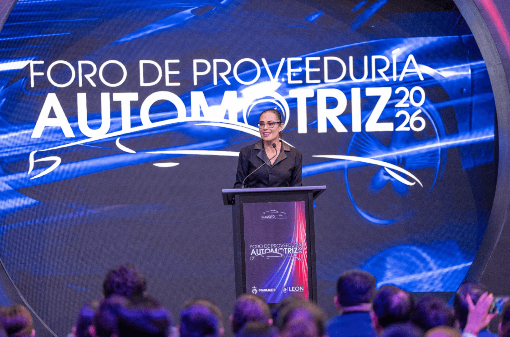 Mujer hablando en foro automotriz en Le&oacute;n, Guanajuato, M&eacute;xico, con p&uacute;blico presente y pantalla digital de fondo.