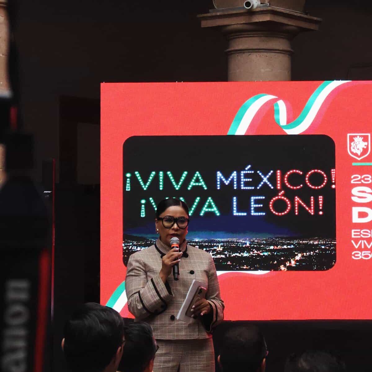 Mujer hablando en evento en León, Guanajuato, con pantalla que dice Viva México en Fiestas Patrias.