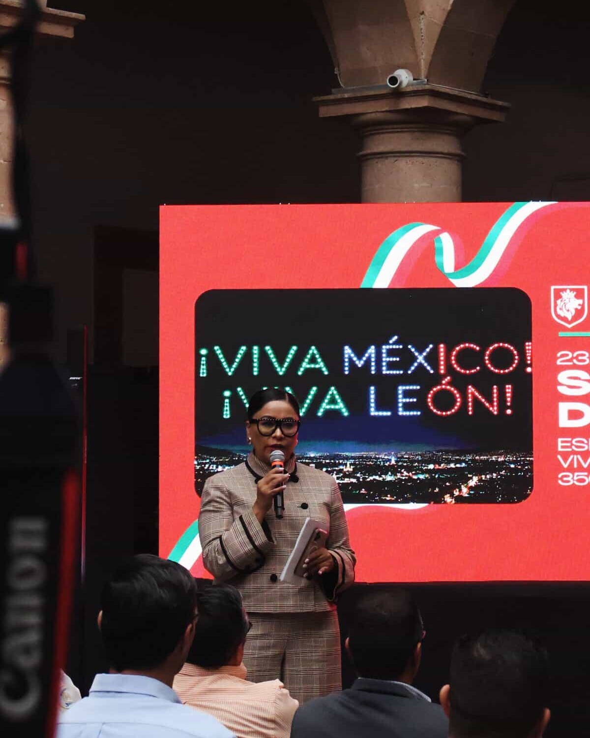 Mujer hablando en evento en León, Guanajuato, con pantalla que dice Viva México en Fiestas Patrias.
