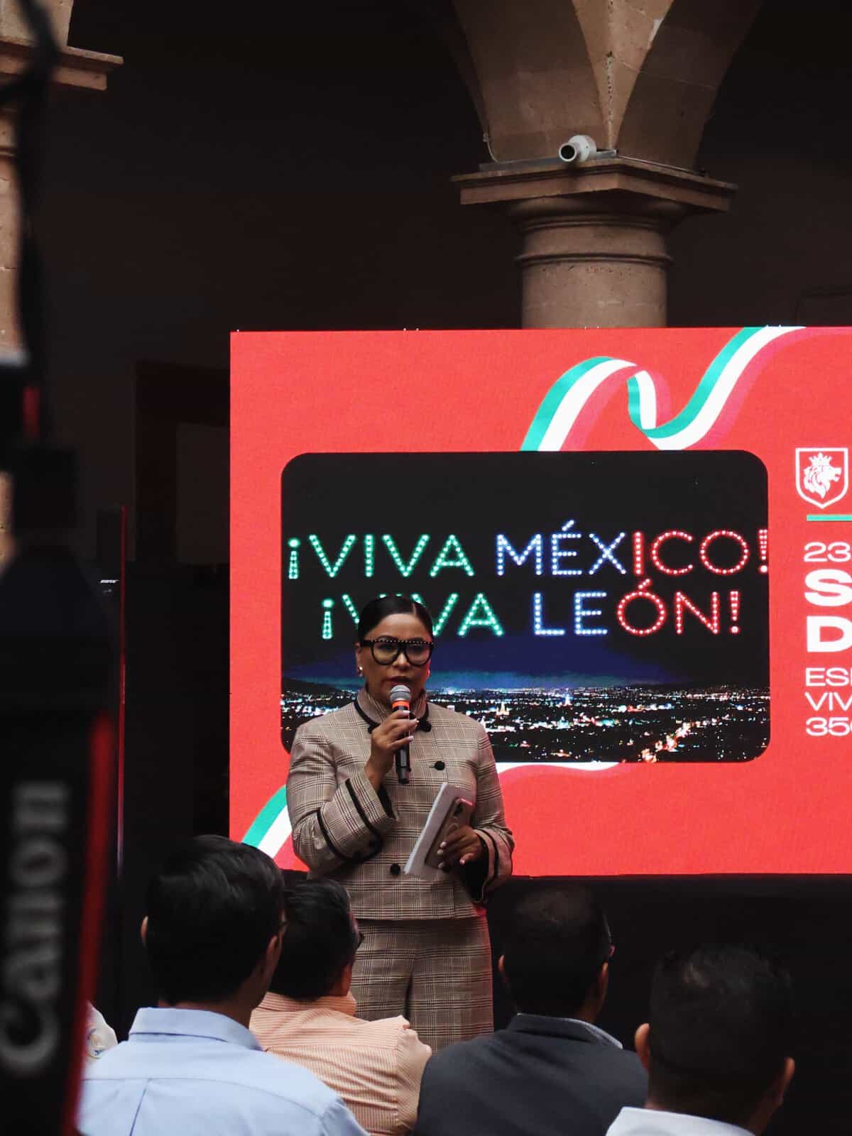 Mujer hablando en evento en León, Guanajuato, con pantalla que dice Viva México en Fiestas Patrias.