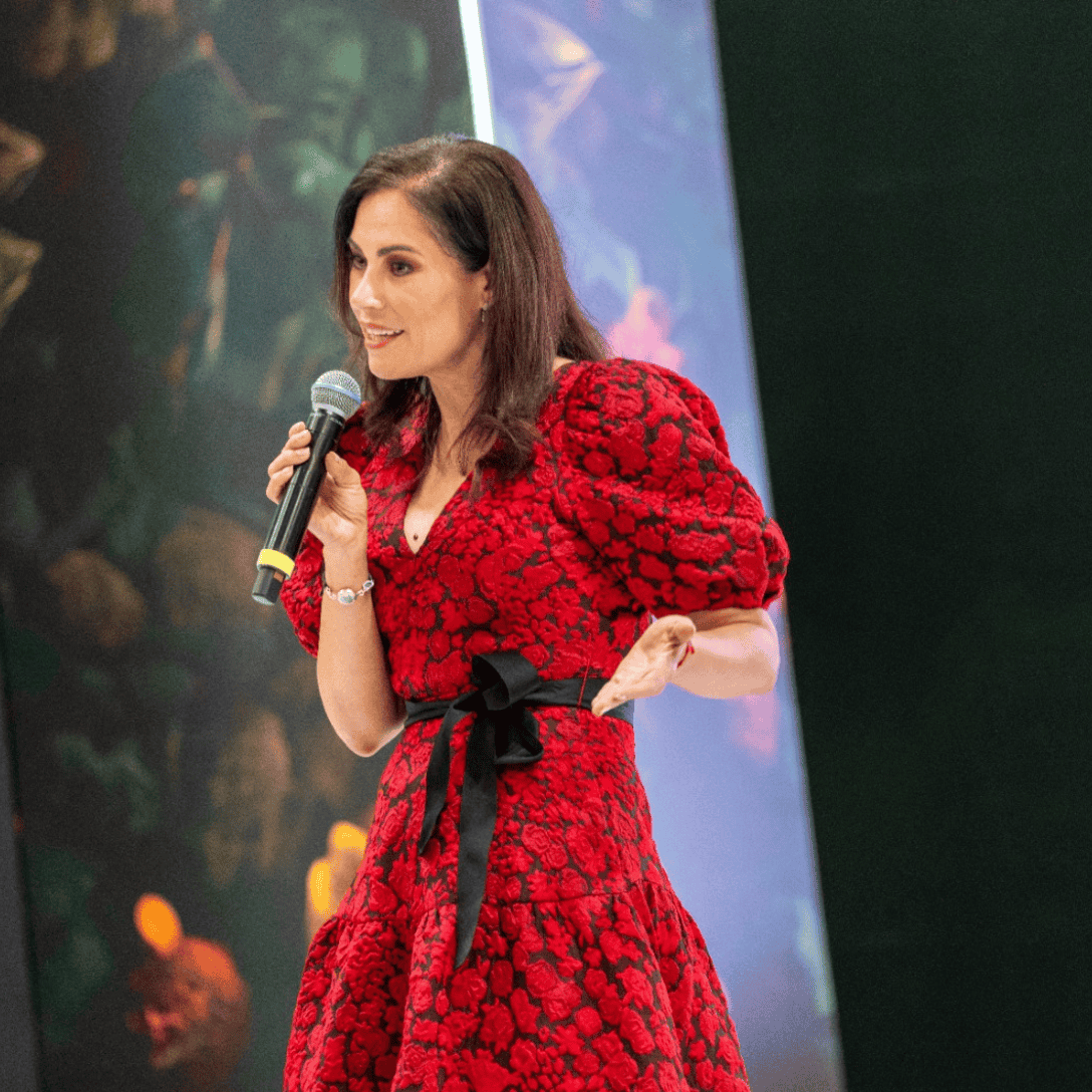 Oradora femenina con micrófono en escenario, vestida con vestido rojo y lazo negro, ante fondo de escenario y pantallas.
