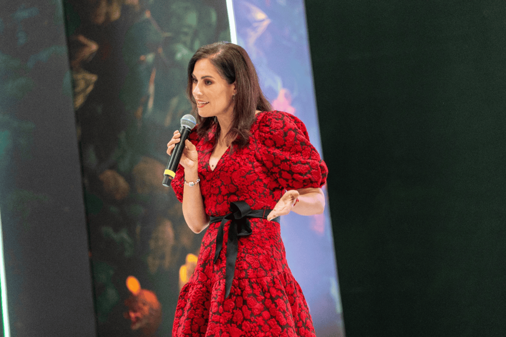 Oradora femenina con micr&oacute;fono en escenario, vestida con vestido rojo y lazo negro, ante fondo de escenario y pantallas.