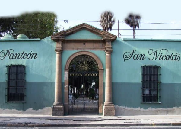 Entrada al Pante&oacute;n San Nicol&aacute;s en Le&oacute;n, Guanajuato, con muro azul y puertas de hierro, rodeado de palmeras y &aacute;rboles.